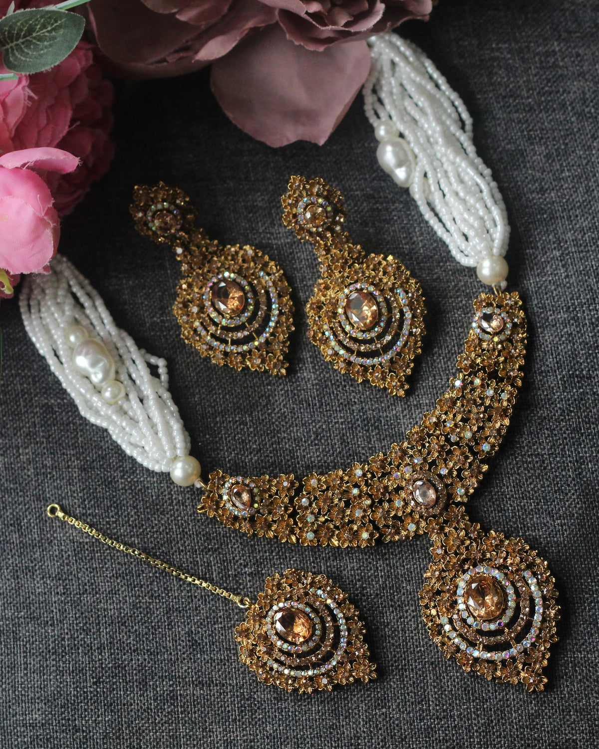 An ornate gold-toned traditional jewelry set laid flat on a dark textured surface, including a beaded necklace, matching earrings, a maang tikka, and white pearl-bead strands with flowers in the corner.