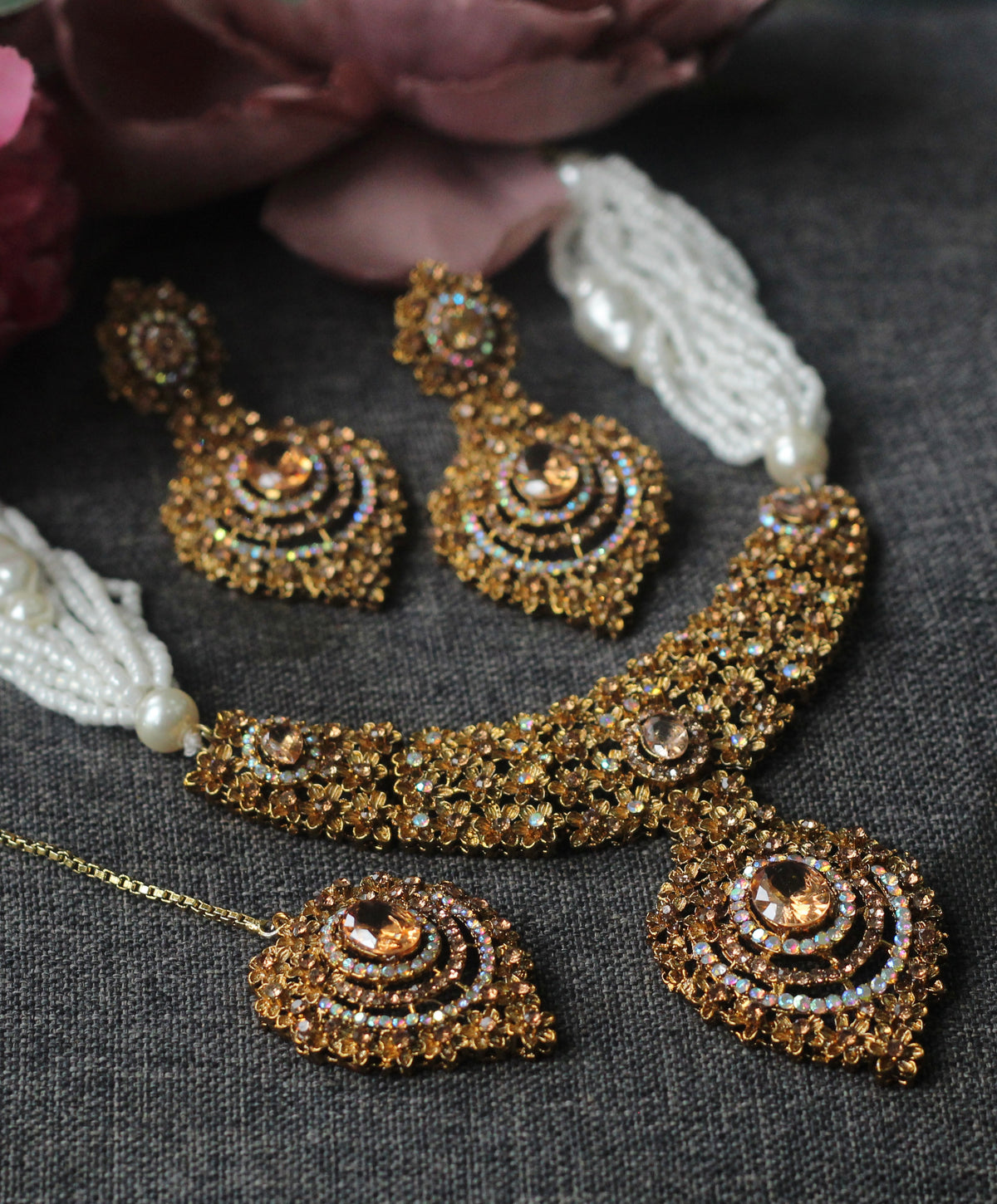 A gold-toned traditional jewelry set including a necklace, earrings, and a matching maang tikka, decorated with intricate crystal work and white pearl tassels, displayed on a dark gray textured fabric.