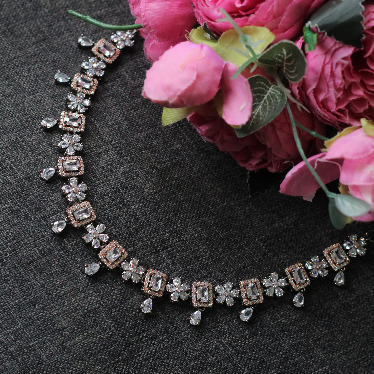 A diamond and rose gold necklace with floral and rectangular motifs, styled on a dark gray textured fabric next to pink roses.